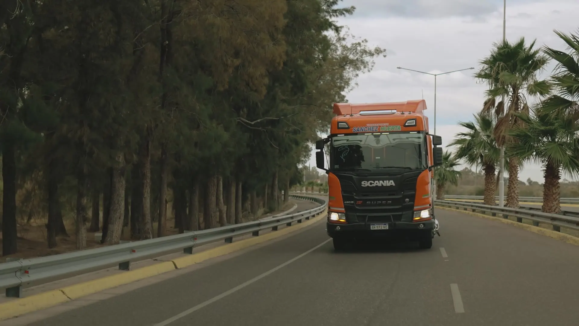 Camión Scania naranja de Transporte Sanchez Huerta con las luces encendidas, avanzando por una autovía entre pinos y palmeras