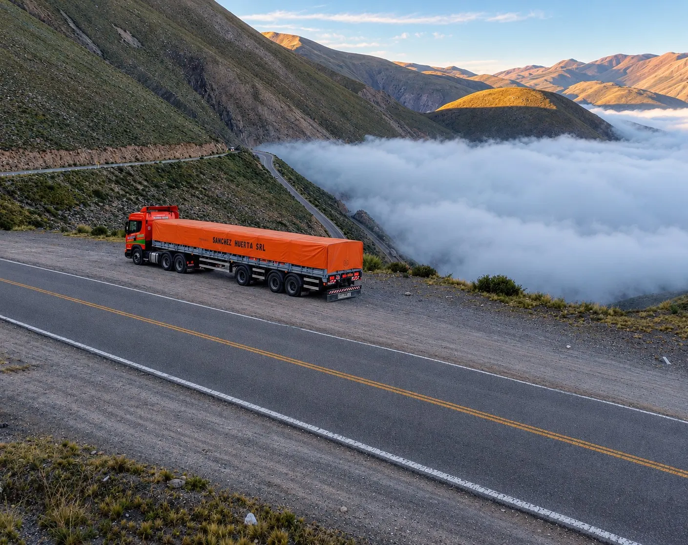 Camión Scania naranja de Transporte Sanchez Huerta con semirremolque de lona detenido al costado de una ruta de montaña, sobre un valle cubierto por un mar de nubes