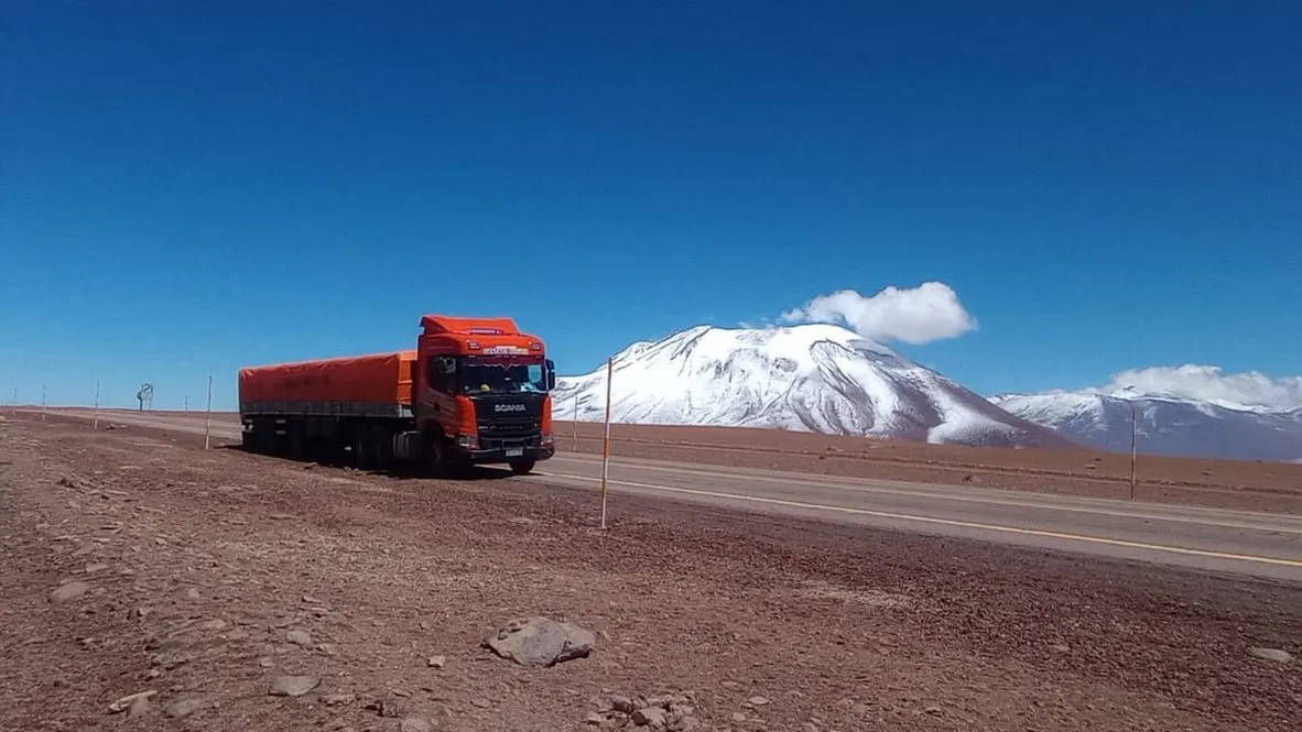 Camión con semirremolque de lona naranja en una llanura de altura de tierra rojiza, con un volcán nevado en el horizonte