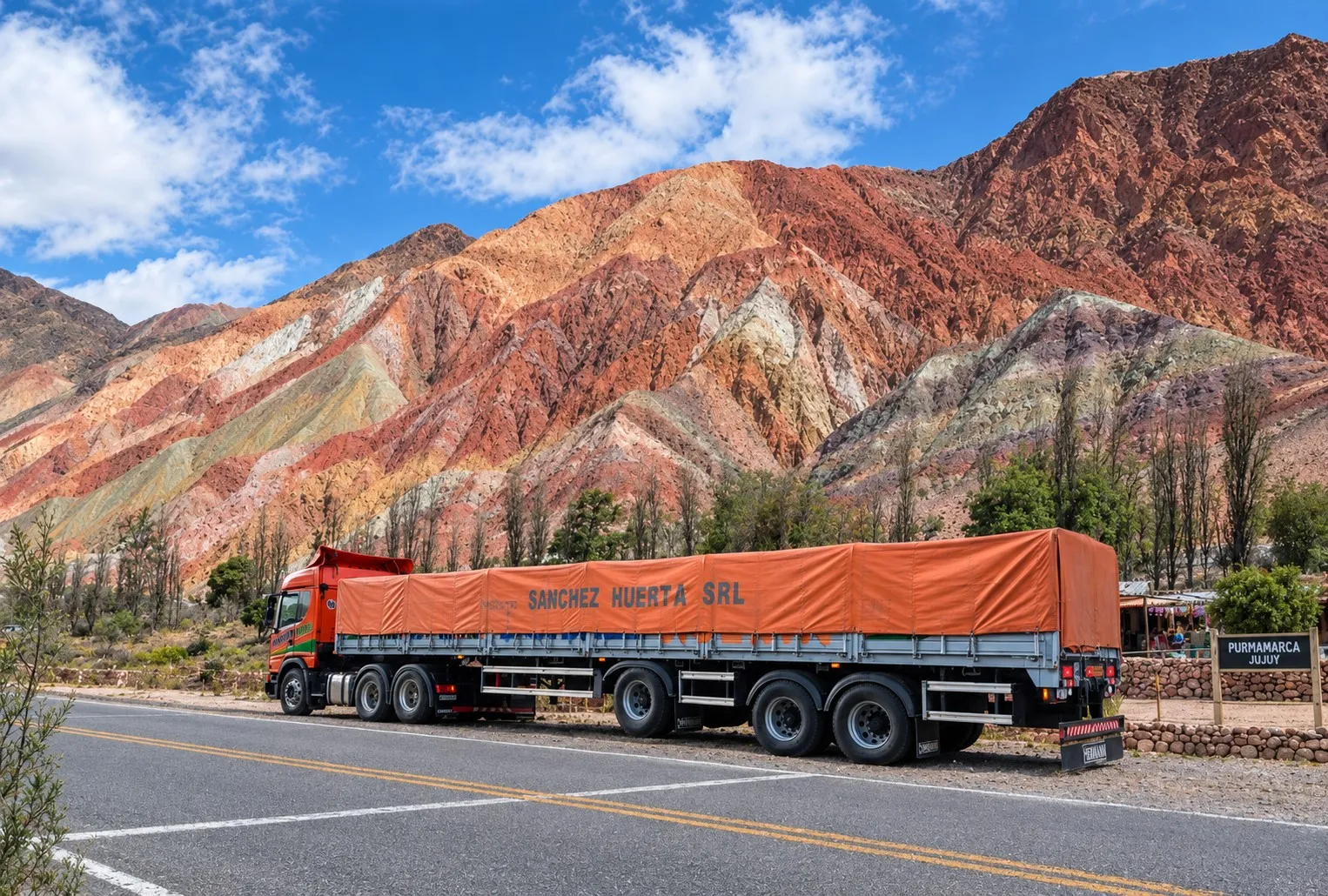 Camión Scania de Transporte Sanchez Huerta con semirremolque de lona naranja en la ruta frente al Cerro de los Siete Colores, junto al cartel de Purmamarca, Jujuy