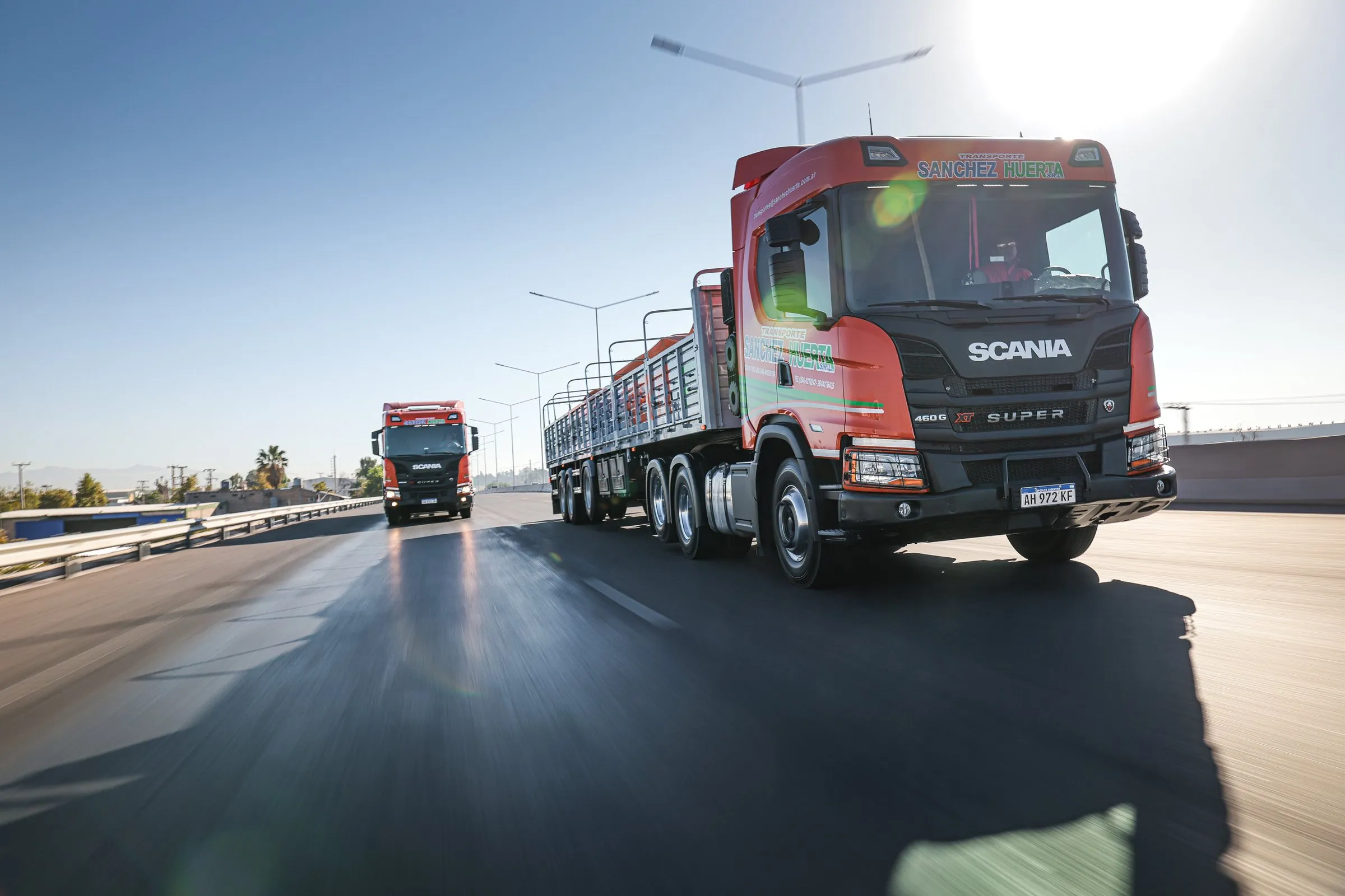Dos camiones Scania naranja de Transporte Sanchez Huerta en fila por una autopista, el primero en primer plano, con la ruta barrida por el movimiento