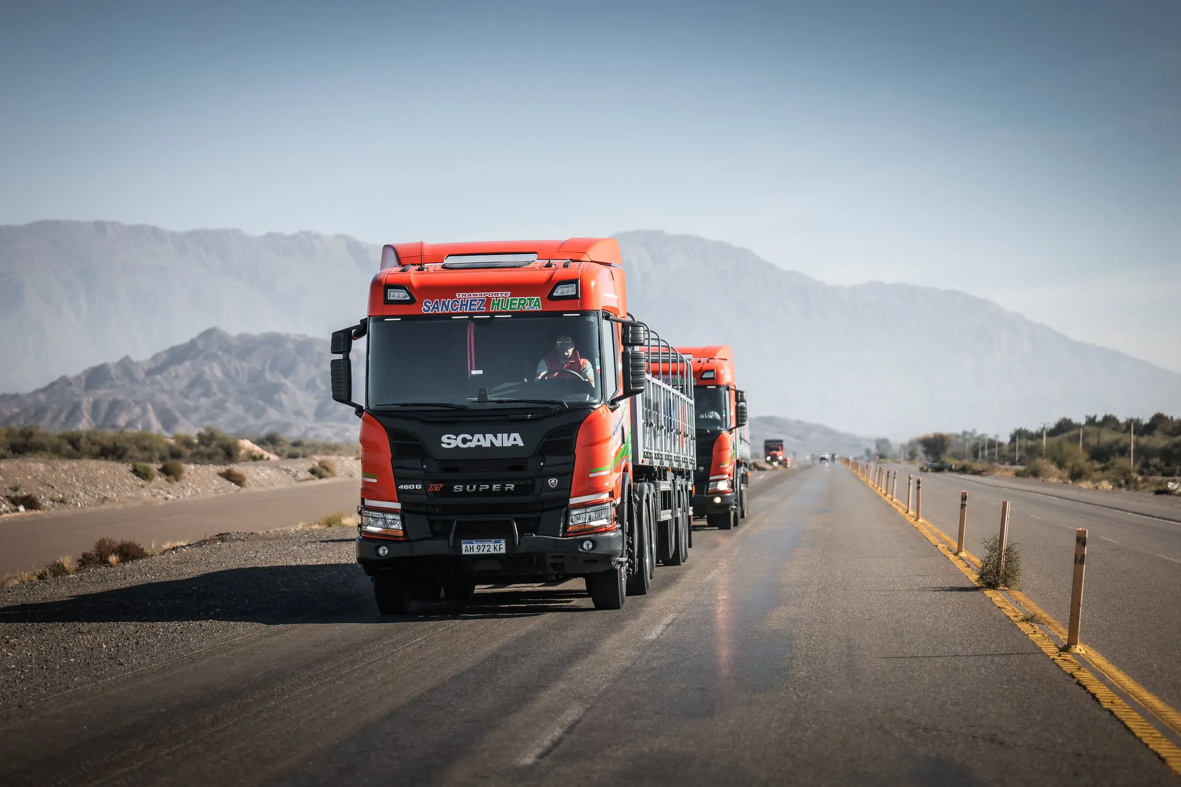 Camión Scania naranja de Transporte Sanchez Huerta con semirremolque de barandas avanzando por una ruta recta, con la precordillera de San Juan de fondo