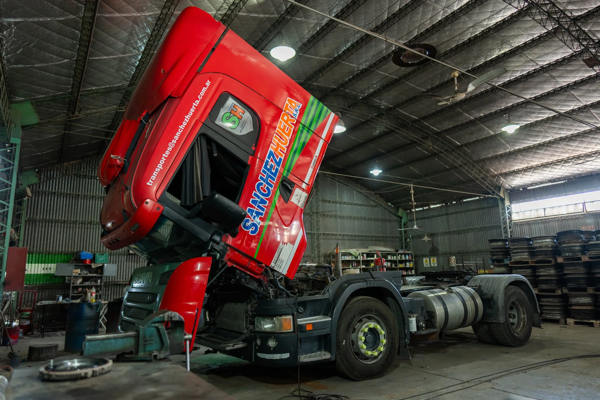 Tractor Scania de Transporte Sanchez Huerta con la cabina levantada para mantenimiento dentro del taller propio, con llantas apiladas al fondo